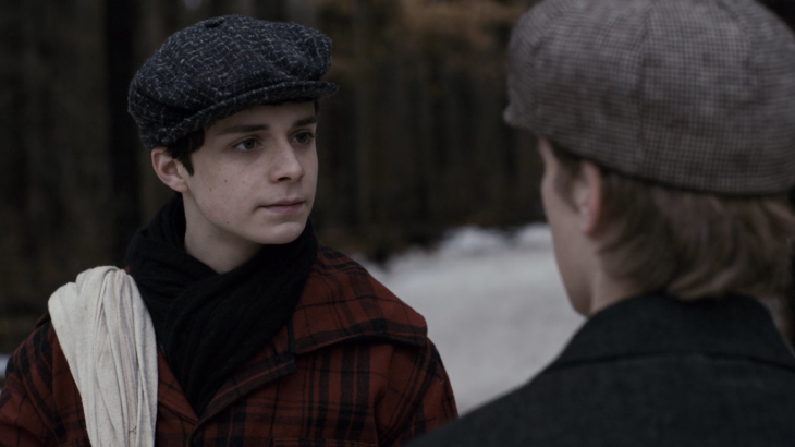 Gilbert looks at another boy. He is wearing a beret, a red plaid coat, and a neatly tucked black scarf.