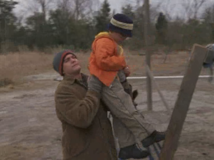 Medium shot of Pacey, wearing outdoor clothes and hat, lifting a tiny boy, also in outdoor clothes and hat, off a ladder.