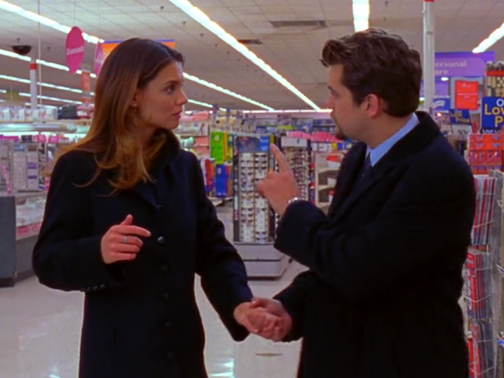 Joey (left, in a black coat) and Pacey (right, in a black coat) are inside a large store, looking at each other, and holding hands. Pacey's hand is