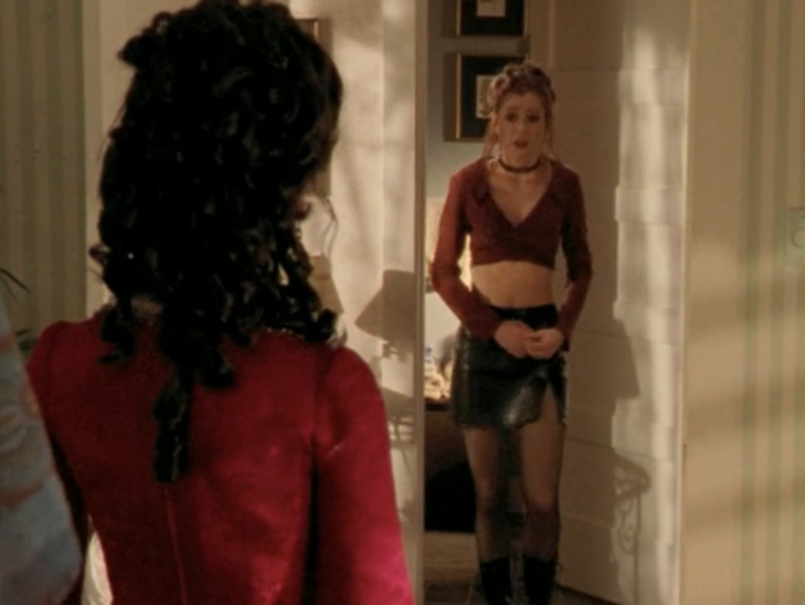 Willow stands in front of a doorway wearing a choker, a messy bun, a red crop top, a leather skirt, and sheer black tights.